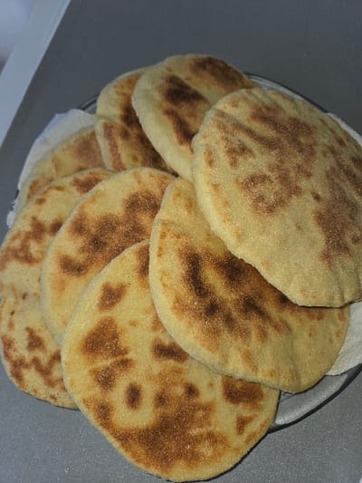 In Blainville-sur-Orne, France, a still-life scene depicts a stack of approximately ten to twelve round, light yellowish-brown flatbreads. Each flatbread exhibits irregular, darker brown, pan-fried or griddle-cooked markings across its surface. The flatbreads are arranged on a white paper napkin, which rests inside a clear, shallow dish or plate, likely glass or plastic. This plate is situated on a smooth, flat, gray surface, possibly a kitchen countertop or table. A lighter gray, undefined background element is visible in the upper left portion of the image. No individuals or active interactions are present.