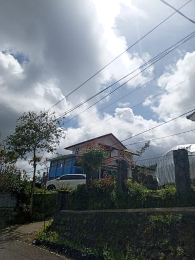 This daytime photograph captures a residential scene in Hinabangan, Philippines, viewed from a street looking upwards towards a house situated on an elevated plot of land. The upper portion of the frame is dominated by a bright, partially cloudy sky, where sunlight breaks through white and grey clouds. Several utility wires crisscross the sky, adding an element of urban infrastructure.

In the midground, a multi-story house features a prominent white roof and blue-painted sections on its lower floors. A light-colored vehicle, appearing to be a white SUV or minivan, is parked directly in front of the house's blue section. The property is surrounded by dense, lush vegetation, including trees with green foliage on the left and various shrubs and plants, some displaying reddish leaves or flowers, around the house. Stone retaining walls delineate the property's boundary in the foreground and midground. To the far right, a part of another structure, possibly a covered area or greenhouse with a mesh fence, is visible.

No people are present, indicating a quiet, everyday moment. The time of day appears to be bright, with strong sunlight despite the cloud cover, suggesting either late morning or early afternoon. No visible text is discernible in the image.