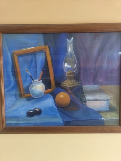 This is a painting framed in wood, depicting a still life. The background features draped blue and purple fabric. In the foreground, on a blue cloth, are several objects. To the left, a small white vase holds paintbrushes. Next to the vase is a wooden picture frame. In front of these items are two dark, oval-shaped objects, possibly olives or dates. To the right of the vase is a round, orange object, likely a fruit. Further to the right, a glass oil lamp with a brass base stands on a dark surface. To the right of the lamp are two stacked books. The painting is hung on a beige wall. The painting's location in Priobskiy, Russia, is not discernable from the image itself.