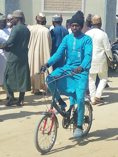 A young adult male, wearing a teal tunic and matching trousers, a black knit cap, black socks, and gray slip-on shoes, is positioned on a black bicycle with red front forks. His hands are on the handlebars, and his head is turned slightly to his left. The bicycle features silver wheel rims and a visible rear suspension component. He is situated on an unpaved, light brown ground surface. Behind him, several adult males are gathered, some in long traditional robes of dark green, beige, and white, with assorted skullcaps. One individual on the far left wears a white face mask. A light-colored building with brown brick-like detailing is visible in the background, along with a partial view of a motorcycle on the right. The scene is outdoors under bright daylight, casting clear shadows. This scene is located in Darmanawa, Nigeria.