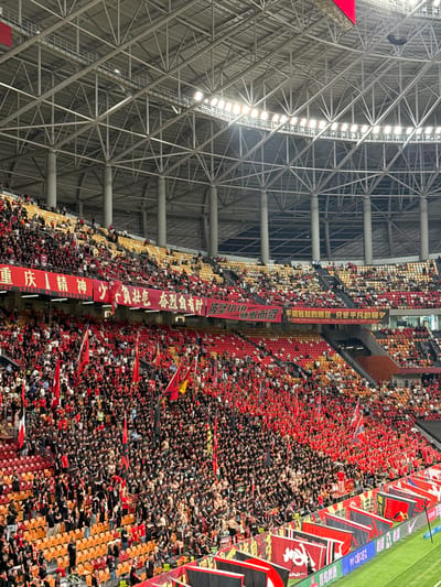 The image captures a densely packed stadium in Longxing, China, with tiers of spectators filling the seats. The majority of the crowd is dressed in black and red, some waving flags. A large red banner with Chinese characters is draped vertically, along with other black and red banners with similar characters, likely representing team slogans or messages of support. The stadium's architecture is visible, featuring a prominent steel grid structure on the left side and bright lighting. Some green grass appears in the upper right corner of the image.