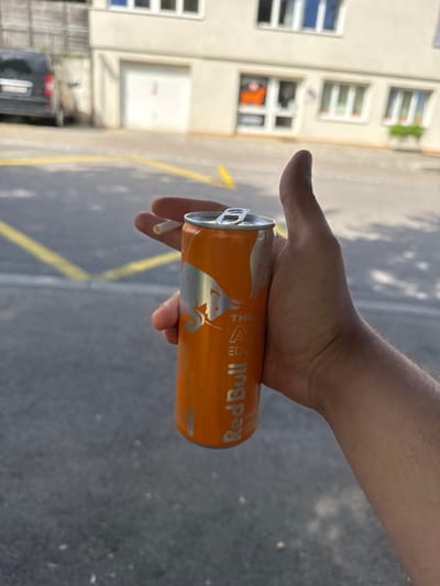 An outdoor shot depicts a person's hand holding an orange Red Bull can and a cigarette. The hand is positioned centrally, gripping the orange Red Bull can which features silver lettering spelling out "Red Bull" and part of "THE EDITION," along with the brand's iconic bull logo. The can's pull-tab is open. A white cigarette is held between two fingers of the same hand.
The background reveals a sunny daytime scene. Bright sunlight casts distinct shadows on the paved ground, which has yellow markings, possibly indicating a parking area or street. To the left, a multi-story, light-colored building with several windows is visible. An orange item, possibly a garment, can be seen hanging in one of the lower windows. A dark vehicle is partially visible parked near the building in the bottom left of the frame. The setting is in Lausen, Switzerland.