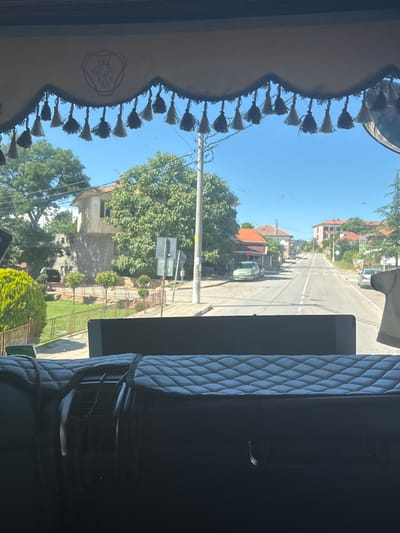 This image appears to be taken from inside a vehicle, looking out through the front windshield. The perspective is from a driver or passenger, with a decorative valance and tassels visible along the top of the windshield.

The scene outside is a rural or suburban street in Mladinovo, Bulgaria, on a clear, sunny day. The street is paved with white lines indicating lanes. Several houses with red-tiled roofs line the street, interspersed with lush green trees. A light-colored car is visible parked on the side of the road, and further down, a green tractor is parked in front of a building. Power lines and poles are also visible, indicating infrastructure. The sky is a bright, cloudless blue, suggesting midday.

To the right of the image, the interior of the vehicle is visible, showing a dark, upholstered seat with a diamond-stitched pattern. The overall impression is one of a journey through a quiet, sunny Bulgarian village. No people are clearly visible. There is no discernible text in the image.