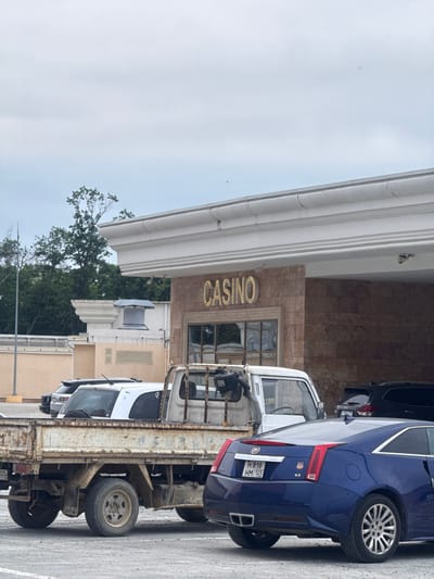 A blue Cadillac sedan is parked in front of a building with a sign that says "CASINO" in gold lettering. The building appears to be a casino, with a marble facade and large windows. A flatbed truck is parked next to the Cadillac, and other cars are visible in the background. The scene is outdoors, and the weather appears to be overcast. The time of day is unclear, but it is likely daytime. The location is Shkotovo, Russia.