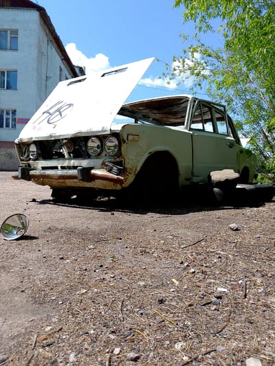 An abandoned, light-colored sedan sits on a gravel and dirt lot under a bright blue sky. The car's hood is propped open, revealing a rusty engine compartment. The vehicle is heavily weathered, with visible rust spots and peeling paint. One of the headlights is missing, and the front bumper is damaged. A large, circular reflector lies on the ground in front of the car. To the right of the car, a spare tire and a large rock are placed, seemingly to prop up the vehicle. In the background, a light blue, multi-story building with visible windows stands to the left, and lush green foliage from a tree drapes over the top right of the car and into the frame. The scene appears to be located in Neryungri, Russia, suggested by context.