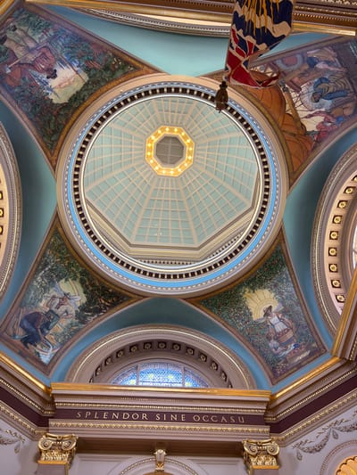 The image shows the interior of the dome of the Legislative Assembly of British Columbia in Victoria, Canada. The dome is octagonal and features a skylight at its apex, which is illuminated. The interior is richly decorated with murals, gold accents, and a central inscription.
The murals depict scenes of early British Columbia, including images of people working in forests and harvesting fruit. These murals are set within arched sections of the dome.
The inscription on a frieze reads "SPLENDOR SINE OCCASU," which is Latin for "Splendor Without Setting." The frieze is above a stained-glass window.
A Canadian flag, partially obscured, hangs from the ceiling in the upper right portion of the image. The overall impression is one of grandeur and historical significance.