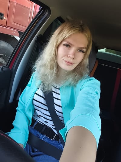 A young woman with blonde, wavy hair is sitting in the driver's seat of a vehicle, looking at the camera and smiling slightly. She is wearing a black and white striped shirt, a bright turquoise blazer, blue jeans, and a black belt. A seatbelt is fastened across her body. The interior of the car is visible, including a portion of the dashboard and what appears to be another vehicle outside the driver's side window. The scene is consistent with a selfie taken inside a vehicle. There is no specific indication of the city of Ягул, Russia.