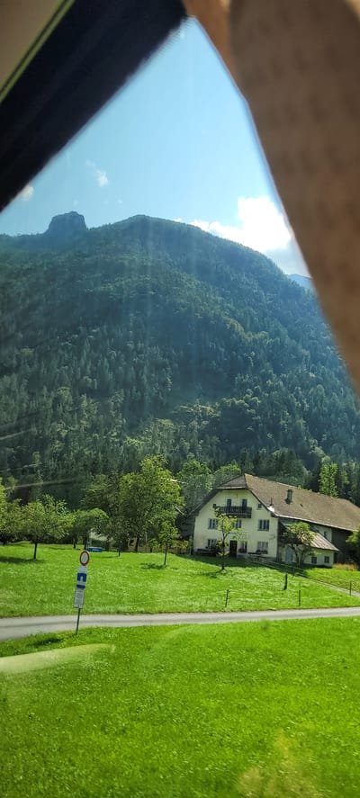 In a bright, sunlit outdoor scene, a forested mountain dominates the background under a clear blue sky with scattered white clouds. The mountain's slope is densely covered with evergreen trees, with a prominent, rocky peak visible at the summit. In the mid-ground, a traditional Austrian farmhouse with a light-colored facade, dark-tiled roof, and balconies stands on a lush green lawn. Several mature trees surround the house. A paved road curves through the lawn in front of the house, and a road sign with a circular red and white symbol and a rectangular blue sign below it is placed on the side of the road. The foreground consists of a vibrant green, well-maintained lawn, suggesting a rural or semi-rural setting. The image is partially obscured by a dark-colored frame in the upper left and a blurred human arm in the upper right, indicating the photo was taken from inside a vehicle or through a window. The location is identified as Georgenberg, Austria.