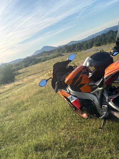 A motorcycle helmet rests on the gas tank of an orange and white sportbike parked on a grassy hill. The motorcycle is facing right, and its rear is slightly angled towards the viewer. In the background, rolling hills covered in green trees stretch towards a hazy mountain range under a blue sky with wispy white clouds. The sun is low in the sky, casting a warm glow. The location is the village of Gigintsi, Bulgaria.