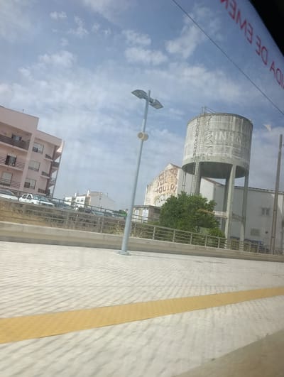 In the city of Pinhal Novo, Portugal, an outdoor scene is captured, likely from a moving vehicle, with a clear sky featuring scattered white clouds. In the foreground, a textured paving stone surface with a yellow tactile warning strip runs horizontally. A tall, cylindrical concrete water tower stands prominently, supported by multiple legs, with graffiti visible on its surface. Next to the water tower, a building displays faded lettering. To the left, a light-colored apartment building with balconies is visible, with several cars parked in front. A lamppost with a light fixture is positioned in the middle ground, and a metal railing lines the edge of an elevated area. The overall impression is an urban environment under a bright, sunny sky.