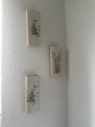 Three rectangular ceramic tiles are mounted on a textured white wall, possibly in a home in Bad Blankenburg, Germany. The tiles are arranged in a staggered formation, with one tile positioned above and between the other two.
The top tile features a painted illustration of dried grass or reeds, depicted in shades of brown and green, with delicate seed heads. The bottom left tile displays a floral arrangement with pink, yellow, and white flowers, along with green foliage. The bottom right tile has a similar floral design, featuring pink flowers and greenery. All three tiles have a light, off-white background. The wall itself has a rough, stucco-like texture. The lighting suggests an indoor setting, possibly during daylight hours, given the soft illumination and lack of strong shadows. No text is visible in the image.