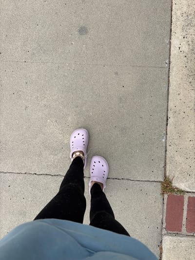 The image shows a first-person perspective of someone standing on a concrete sidewalk in Seal Beach, California. The person is wearing black leggings and lavender Crocs. A blue sweatshirt partially obscures the right side of the frame. The sidewalk is cracked in places and abuts a different colored section of concrete and a small area with red brick. There are some small weeds growing in the cracks near the brick area.