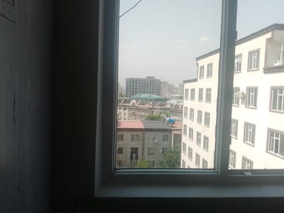 The image, taken from an interior vantage point, looks out through a window onto a cityscape. The window frame, partially visible on the left and in the center of the image, is a light color and appears to be made of a composite material.
The view outside reveals a dense urban environment with multiple buildings of varying heights and architectural styles. In the mid-ground, several multi-story apartment buildings are visible. Some have light-colored facades with numerous windows, while others appear older, with more uniform designs. One building in the mid-ground has a distinctive red and dark gray roof. Satellite dishes are mounted on the roofs of several buildings.
Further in the background, taller, more modern buildings can be seen, suggesting a developing urban area. The sky is overcast or hazy, creating a muted light. Trees are interspersed among the buildings, indicating some green spaces within the city. The overall impression is of a residential and commercial district.
Considering the context provided, this scene depicts a view in the city of Vahdat, Tajikistan. The architecture and urban density are consistent with such a location. There are no people or specific actions clearly discernible in the image. The focus is on the built environment and the urban landscape.