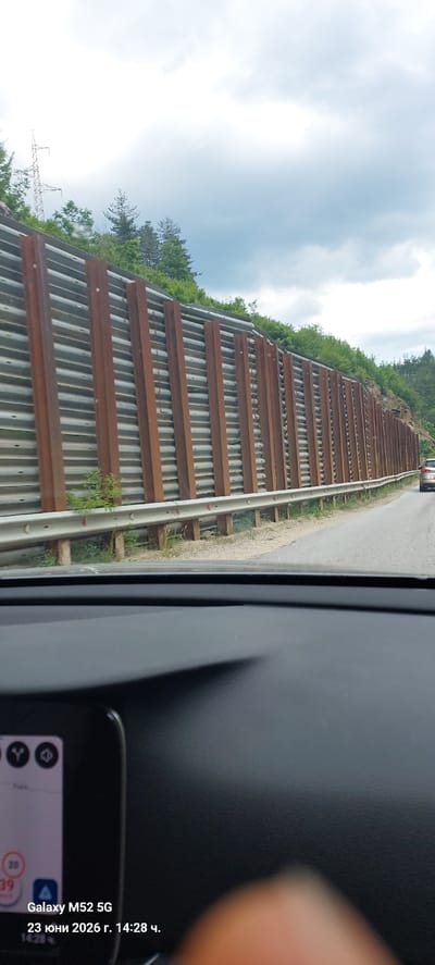 The image shows a view from inside a vehicle looking out at a road with a sound barrier along the side. The sound barrier is made of corrugated metal sheets supported by vertical wooden posts that appear weathered and rusted. Green trees and foliage are visible on the hillside behind the barrier. A silver car is driving ahead on the road. The dashboard of the vehicle is partially visible in the foreground, with a portion of a smartphone screen displaying the text "Galaxy M52 5G" and "23 юни 2026 г. 14:28 ч." This indicates the photo was taken on June 23, 2026, at 2:28 PM. The overall environment suggests a rural or mountainous road. While the exact location in Laka, Bulgaria, is not visually identifiable from the image alone, the presence of a road, sound barrier, and natural landscape is consistent with such an area.