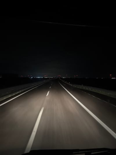 The image depicts a nighttime scene from the interior of a moving vehicle on a multi-lane highway, located near Cerignola, Foggia province, Italy. The environment is dark, with a clear or lightly clouded sky. The asphalt road surface is illuminated by the vehicle's headlights, revealing two lanes of travel in the foreground, separated by a dashed white line. Solid white lines mark the outer edges of the carriageway. Metal guardrails are visible along both sides of the road. In the far distance, a horizontal array of artificial lights is present, consisting of diffuse clusters of white/yellow lights indicative of a distant town or industrial area, interspersed with multiple distinct red obstruction lights, consistent with active wind turbines common in the region. No individuals are visible within the frame. The scene captures the act of nocturnal travel along the highway.