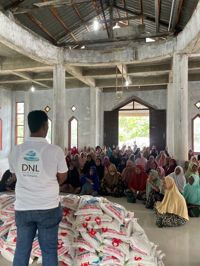 The image depicts an indoor community gathering at Meunasah Ara, Indonesia, likely for a distribution event. In the foreground, a man wearing a white t-shirt with the text "DNL body & bone for humanity" and a circular logo stands with his back to the camera. To the right, numerous white sacks are stacked, bearing text such as "BRUTTO: 5 Kg", "KUALITAS TERJAMIN" (Guaranteed Quality), "KILANG PADI", "ACEH RICE SPECIAL", "IKU PAYA", and "Beras Tangse", along with product codes like "JDM001211076" and "IDM001211076", indicating they contain rice.
Beyond the sacks, a large group of people, primarily women and youth, are seated on the concrete floor, facing towards the front of the room. Many of the women are wearing headscarves (hijabs) in various colors. The setting is a simple building with concrete pillars, plain concrete walls, and an exposed roof structure featuring corrugated metal sheets and wooden beams. Natural light streams in through several windows and a large doorway at the back, suggesting it is daytime. Fluorescent strip lights are mounted on the ceiling beams, and a small ceiling fan is also visible. The scene captures an organized event focused on providing aid or resources to the local community.