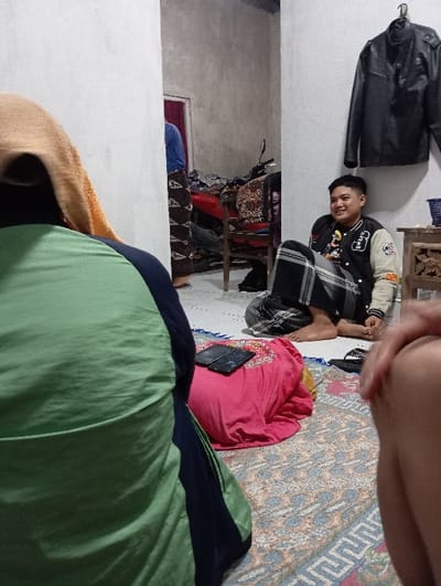 The image portrays an indoor scene within a residence in Bulukandang, Indonesia. In the immediate foreground, on the left, the back of an individual is visible, wearing a light green top over a dark blue garment and a tan head covering. To the right, a bare leg and thigh are partially seen, belonging to a person positioned close to the camera.
In the mid-ground, a young male with short dark hair, wearing a black and white varsity jacket and patterned dark trousers, is seated directly on a patterned rug. He is smiling and looking towards the left. His bare feet are visible, with a single black slipper resting nearby. Behind the foreground left individual, a folded pink garment or blanket is situated on a blue mat, with a dark smartphone resting on top of it.
The floor is covered by a patterned rug featuring geometric designs in blue, orange, and white tones. The walls are light-colored, appearing to be unfinished or lightly painted concrete. A black leather jacket hangs from a hook on the wall to the right. Adjacent to the seated male, a small carved wooden table holds a dark blue basket or container.
In the background, a person wearing a blue top and dark patterned trousers is partially visible, standing near a red motorcycle or scooter. A wooden chair draped with a patterned cloth is positioned near the motorcycle. A dark red door frame is visible in the distant background. The lighting is moderate, typical of an indoor environment.