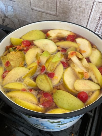 In Pervomaisk, Ukraine, a close-up, overhead shot captures a cooking pot filled with sliced apples and what appear to be cherries or small red berries, submerged in a simmering liquid. The pot, white with a black rim, sits on a black stovetop. The background features a tiled wall with a subtle pattern, suggesting a kitchen environment. The apples are cut into various shapes, some retaining their core, and are a mix of green and yellow. The red berries are small and round, interspersed among the apple slices. Bubbles on the surface of the liquid indicate that the contents are actively cooking, likely on a low heat. The overall scene depicts food preparation in a domestic setting.