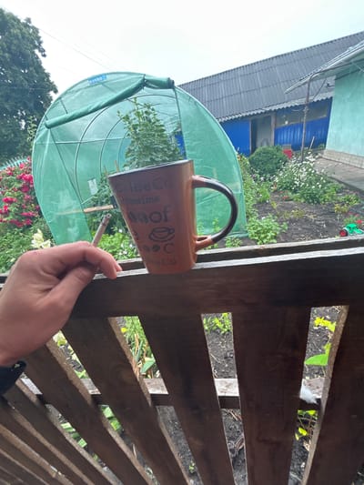 A close-up shot focuses on a person's hand holding a cigarette and a coffee mug. The hand is positioned over a wooden railing, with the mug to the right of the cigarette. The mug is brown with a dark brown handle and features text and images related to coffee. In the background, a green greenhouse is visible, along with a garden containing flowers and plants. A blue-walled building with a corrugated roof stands behind the greenhouse. The scene appears to be an outdoor setting, possibly a backyard in the village of Terny, Ukraine. The overall atmosphere is casual and domestic.