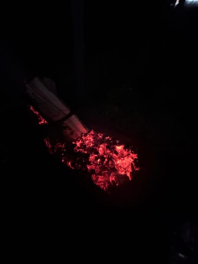 The image depicts a scene at night in the city of Мишкино, Russia, dominated by a source of intense red light from glowing embers and partially burned wood. A substantial cluster of bright red and orange embers occupies the lower and middle-right portions of the frame. Above and slightly to the left of the main ember bed, a thick, partially consumed log or piece of wood is angled diagonally from the upper left towards the lower right. Sections of this log, particularly where it meets the embers, are visibly glowing red. A dark, band-like element, possibly a strap or unburnt portion, is wrapped around the log. The vast majority of the image beyond the immediate vicinity of the fire is completely dark, indicating an outdoor, nighttime environment with no discernible background features or individuals present.