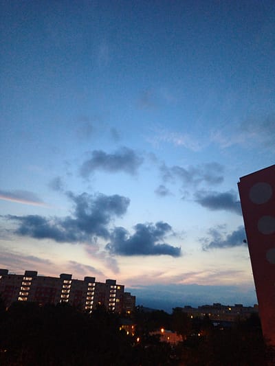 The image is a long shot of a city skyline at dusk. The sky is a gradient of blue, transitioning to lighter shades near the horizon. Several dark, scattered clouds are present in the sky. In the foreground, silhouetted trees and foliage obscure the ground. To the left, a long, multi-story apartment building with many lit windows is visible, its facade appearing to be light brown or orange. In the middle ground, another apartment building or complex with fewer lit windows is seen. To the right, a portion of a red building with large white circular patterns is in the frame, extending from the right edge upwards. In the background, more distant buildings with some lit windows are faintly visible, along with streetlights. The scene suggests a residential area in the city of Most, Czechia, at twilight.