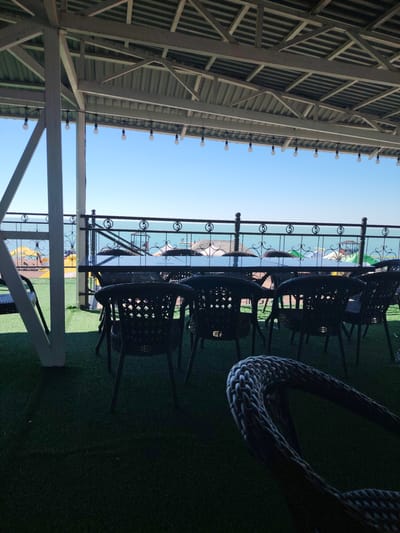 The image shows an outdoor seating area at a cafe or restaurant on the shore of Lake Balkhash in Toranghylyq, Kazakhstan. The foreground features dark wicker chairs and a table, with a braided rope chair element in the lower right corner. The seating area is situated on a green artificial turf surface, possibly Astroturf. Beyond the seating, a black metal railing with decorative scrollwork separates the area from the lake. The lake's water is a vibrant turquoise, and the sky is a clear, pale blue. In the distance, across the lake, beachgoers can be seen with yellow umbrellas and inflatable watercraft, indicating a recreational area. The seating area is covered by a light-colored, slatted roof structure, supported by white pillars. A string of small, white globe lights is suspended from the roof. The overall impression is of a relaxed, seaside dining or lounging establishment.