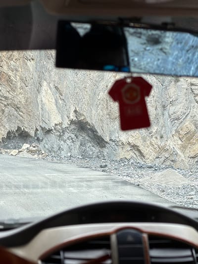 A first-person perspective from inside a vehicle shows a rough, gray asphalt road winding through a mountainous landscape. The road is flanked by a steep, rocky cliff face composed of layered gray and tan stone. Loose rocks and scree are scattered along the base of the cliff and on the shoulder of the road. In the foreground, the blurred dashboard of the vehicle is visible, including a portion of the steering wheel and center console. Hanging from the rearview mirror is a small, red t-shirt-shaped air freshener with a circular emblem. The scene suggests travel through a remote, rugged, and possibly hazardous terrain. The image location is Stongde, India.