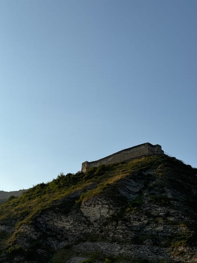 An ancient stone fortress stands atop a grassy, rocky hill under a clear blue sky. The fortress appears to be a long, low structure with a flat roof and stone walls. The hill is covered in green vegetation and has a rugged, uneven surface. In the background, more hills are visible, partially obscured by the trees on the main hill. The lighting suggests it is late afternoon or early evening, casting shadows across the landscape. The image does not contain any people or identifiable human activity. This is a view of the Khuchni Fortress in the village of Khuchni, Russia.