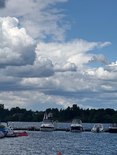 A panoramic, eye-level shot captures a serene waterscape at the Tourist Boarding House "Klyazminskoye Reservoir," Russia. The scene is dominated by a vast expanse of deep blue water, reflecting the dramatic, cloud-filled sky above. A line of boats is docked along a wooden pier in the foreground, their hulls varied in color and size. The largest boat, a white motor yacht with a black canopy, is positioned centrally, its sail furled. To its left, a smaller white boat with a red hull and a white structure is moored. Further to the right, two other motorboats, one with a white canopy and another with a dark hull, are tied up. The background features a dense line of dark green trees, behind which several houses with red roofs are partially visible. The sky is a dynamic mix of bright blue patches and large, fluffy white and gray clouds, suggesting a day with changing weather. The overall impression is one of a peaceful, natural setting on a body of water, with the docked boats indicating a recreational or residential area.