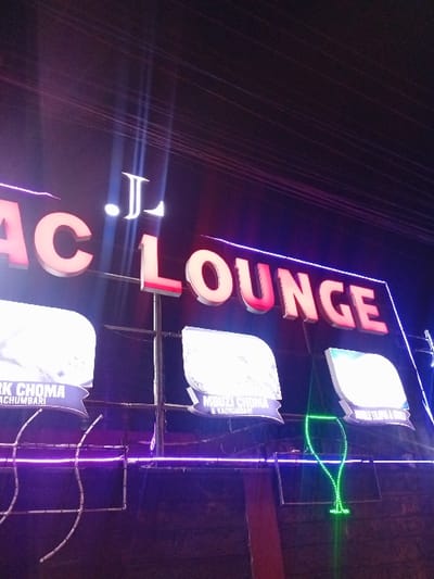 A vertically oriented photograph showcases a neon sign at night. The sign reads "ACI LOUNGE" in bold, illuminated letters. Below the main text are three smaller, lit panels with writing. A green neon light shaped like a cocktail glass is visible. Vertical purple light strips frame the edges of the sign's structure. In the background, darkness is punctuated by electrical wires, and the overall color palette is dominated by vibrant neon hues.