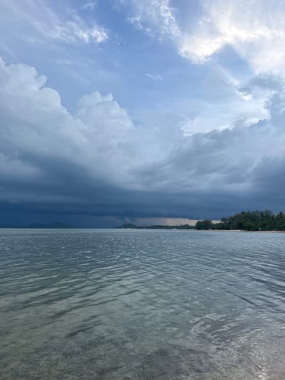 A wide shot shows a body of water with a sandy shore on the right and a treeline in the distance, beneath a dramatic sky. The water is calm, with subtle ripples breaking the surface and showing the sandy bottom beneath. Dark, heavy clouds dominate the sky, with shafts of sunlight breaking through on the upper right, illuminating patches of lighter blue sky. The treeline on the right is dense with green foliage, meeting a sandy beach. In the distance, a few low-lying hills or islands are visible, shrouded in haze and the dark clouds. No people or objects are identifiable in the image. The scene is likely located in Ban Bang Khao, Thailand, given the tropical vegetation and coastal environment.