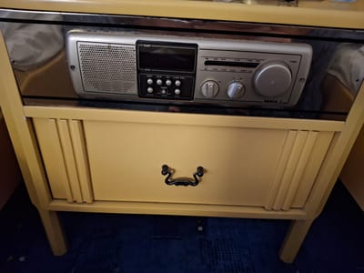 This is a medium shot of a vintage WANDA 2. radio and clock embedded in a light yellow, wood-grain bedside table with a mirrored backsplash. The radio, silver and black, has a digital display that reads "SLEEP" and "AUTO". Below the display are several round buttons labeled "SLEEP", "HOUR", "MIN", "ALARM DISPL", "SNOOZE", "OFF", "BUZ", and "RADIO". To the right of the display is a large tuning dial and a smaller volume dial. The radio has speaker grilles on the left and right sides. The bedside table has a single drawer with a black, ornate metal pull. The setting appears to be indoors, possibly a hotel room, given the furniture style and mirrored surface. The lighting is neutral, suggesting daytime. There are no people visible in the image. The text visible includes the brand name "WANDA 2.", radio band indicators (FM, LW), frequency numbers, and button labels. The image is focused on the radio and the drawer of the bedside table.