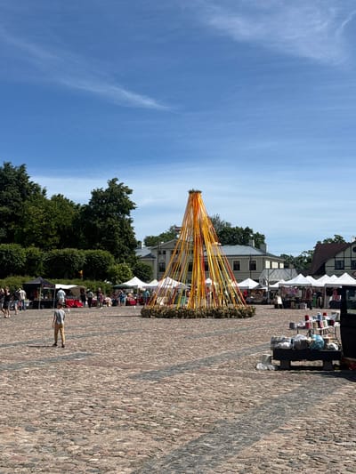 In the city of Dobele, Latvia, a lively outdoor market takes place on a cobblestone square. A prominent, tall maypole adorned with yellow and orange ribbons stands in the center of the square, its ribbons cascading downwards. Around the maypole, numerous white tents are set up, serving as stalls for vendors. People are seen browsing and interacting at these stalls. In the foreground, a young boy walks across the cobblestones, his back to the camera. The environment is sunny with a clear blue sky dotted with wispy clouds. Lush green trees form a backdrop to the scene, with buildings visible behind the tents.