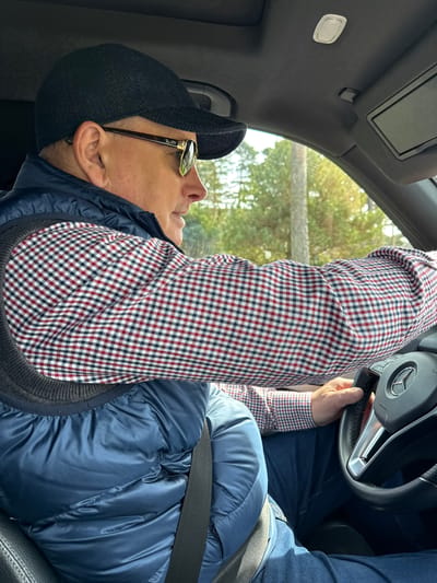 An upright, eye-level, full shot shows a man in the driver's seat of a car. The man, seen from the chest up, is wearing a dark blue puffer vest over a red, white, and blue plaid long-sleeved shirt. He is also wearing dark blue jeans. On his head, he wears a dark grey or black flat cap, and he has gold-rimmed sunglasses with "Ray-Ban" visible on the temple. His head is turned slightly to his right, looking upwards and forward. His left hand rests on the steering wheel.
The car's interior features a black dashboard and headliner, with a sun visor and a small overhead light fixture visible towards the top left of the frame. The steering wheel prominently displays a Mercedes-Benz logo and the word "AIRBAG." A dark seatbelt crosses the man's chest.
Through the car windows, a blurred background of green foliage and what appears to be a light-colored fence or railing is visible, indicating an outdoor setting during daytime. The natural light suggests it is clear or partly cloudy. There are no specific visual cues in the image to confirm the provided location context of Strömstad, Sweden, but the scene is set in a car in that vicinity. The man's expression is obscured by sunglasses, and he appears to be either driving or observing his surroundings while stopped.