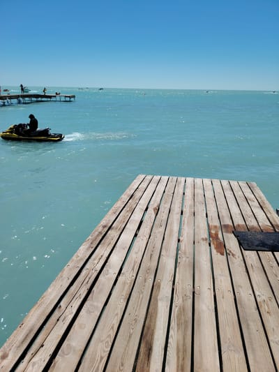 A bright, sunny day over the turquoise waters of the Caspian Sea at Toranghylyq, Kazakhstan. The foreground is dominated by a wooden pier made of weathered planks, running diagonally across the frame from the bottom left to the top right. The planks are rough and have visible knots and nail heads. A small black mat or object is visible on the far right side of the pier.
In the middle ground, a person in silhouette is riding a yellow jet ski, leaving a wake of white spray on the water. The jet ski is moving away from the viewer and towards the left of the frame. Further out in the water, several other jet skis and small boats can be seen, some with people on them.
In the background, a long wooden pier extends from the left side of the frame. On this pier, several figures are visible, including people standing and one person appearing to jump. The horizon is clear and a bright blue sky with no clouds stretches across the top of the image. The overall impression is of a recreational day at the beach.