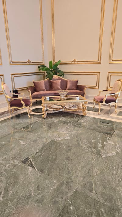 The image shows a luxurious seating area with a brown sofa and two armchairs, placed on a green marble floor. A golden ornate coffee table sits in front of the sofa, adorned with a crystal bowl and two white candles. Behind the furniture, a wall features intricate gold trim paneling. A large green plant stands to the left of the sofa. The scene is likely an interior in a hotel lobby or a well-appointed home in Agios Epiktitos, Northern Cyprus / Cyprus, suggesting a sophisticated and elegant ambiance.