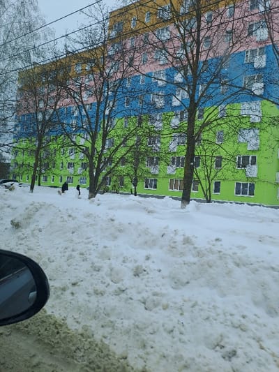 This image captures a winter scene in Sasovo, Russia. The primary subject is a multi-story residential building with a distinct facade featuring horizontal bands of color: yellow-orange at the top, followed by pink, blue, and lime green at the base. The building presents multiple white-framed windows and balconies across its visible levels. Several bare deciduous trees occupy the mid-ground, partially obscuring the structure. The foreground and mid-ground are characterized by significant accumulations of white and greyish snow, forming uneven drifts. Two individuals in dark clothing are observed at the base of the building; one is in a stooped posture, the other stands upright nearby. A dark vehicle is partially visible behind a snowdrift on the far left. The photograph was taken from within a vehicle, with a portion of a side mirror visible in the lower-left corner. The sky is uniformly overcast.