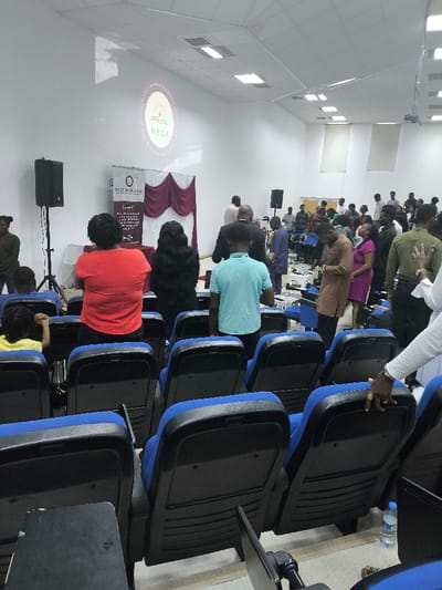 An indoor auditorium in Kızılbaş, Northern Cyprus / Cyprus, hosts a gathering of approximately 30-40 individuals. The room features white walls, a white acoustic tile ceiling with rectangular recessed lighting fixtures, and a light-colored floor. At the front, a circular logo displaying "NECF" with a stylized sun and open book is projected onto the wall. Below this projection, a stage area is defined by a white backdrop with maroon drapes. Two black floor-standing audio speakers on tripods are positioned to the left and right of the stage. In the foreground, rows of blue and black upholstered auditorium seats are visible, some occupied, others empty.
A diverse group of attendees, primarily adults, are present. In the immediate foreground, observed from the rear, are a woman in a red t-shirt, a woman with long dark hair, and a young male in a light blue t-shirt, all standing in an aisle. A young child in a yellow shirt is seated in a front-row seat. Further back, a larger crowd stands in the center and right sections of the room. Attire varies, including casual wear, a brown traditional garment worn by one man, and a purple dress on another individual. Several attendees appear to be engaged, with one person in a green shirt having an arm raised towards the ceiling. A person in white long sleeves is partially visible on the far right, with a hand resting on the back of a seat. A plastic water bottle is on the floor near the right foreground seat. The overall scene suggests a organized event or meeting in progress.