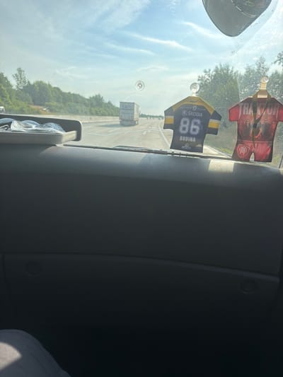 The image shows a view from inside a vehicle, looking out through the windshield. The vehicle appears to be traveling on a highway. In the foreground, on the dashboard, are two novelty air fresheners shaped like sports jerseys. One is black and yellow with the number 86 and the word "ŠKODA" and "BUDINA" visible. The other is red with "TRAKTOR" written on it and an image of several people.

Outside the vehicle, the highway stretches into the distance, with trees and greenery lining the sides. Several trucks are visible on the highway, including one directly ahead in the middle lane. The sky is blue with some scattered clouds. There is a circular suction cup mark on the windshield.

The provided context suggests the location is Hranice, Czechia. Based on this, the scene depicts a typical highway travel experience in that region. No people are clearly identifiable within the vehicle or as pedestrians. The focus is on the interior dashboard elements and the road ahead.