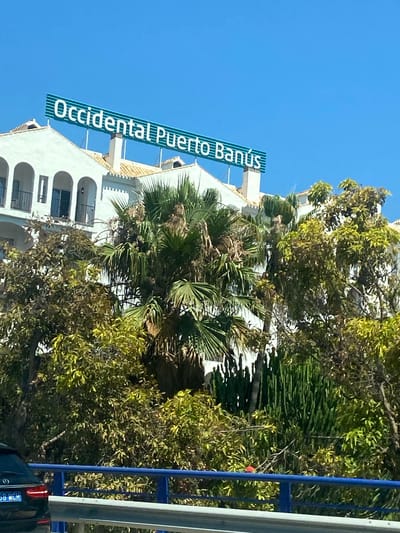 This media file captures a bright, sunny day in Nueva Andalucía, Spain. The scene is dominated by lush green trees and palm fronds, set against a clear blue sky. In the background, a white building with arched windows and a terracotta-tiled roof is visible. A prominent sign on the building reads "Occidental Puerto Banús" in large, white, sans-serif letters against a teal background. The sign is angled, suggesting it is mounted on the side of the building. To the right of the image, a blue metal railing and a white pole are visible, along with a portion of a dark-colored vehicle. The overall impression is one of a warm, Mediterranean environment. The time of day appears to be midday, given the bright sunlight casting distinct shadows and the clear, cloudless sky. There are no people clearly visible in the frame, and no specific activity is taking place beyond the static view of the building and its surroundings. The visual elements suggest a hotel or resort setting, typical of the Puerto Banús area, known for its luxury and scenic beauty. The weather is clearly fair and sunny.