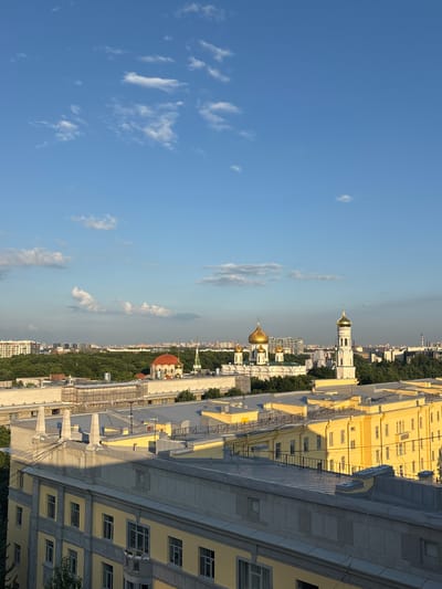This image is a medium-angle, eye-level shot of a city skyline, likely Annino, Russia, under a bright blue sky with scattered white clouds. The foreground is dominated by the grey and yellow rooftops of multi-story buildings. These buildings feature architectural details such as prominent cornices, window frames, and decorative elements on their facades. Some of the rooftops appear to be undergoing construction or renovation, with scaffolding visible on one section.
In the mid-ground, a dense band of lush green trees stretches across the landscape, indicating a significant park or wooded area. Peeking above the trees are several religious structures with golden domes, characteristic of Russian Orthodox churches. The most prominent of these is a white church with multiple golden domes and a separate, taller white bell tower also topped with a golden dome.
The background shows a more distant cityscape with a variety of buildings, some appearing taller and more modern, while others are lower-rise. The overall impression is a blend of residential and religious architecture set against a backdrop of abundant greenery. The lighting suggests it is either early morning or late afternoon, with the sun casting long shadows across the rooftops. No people are visible in the image.