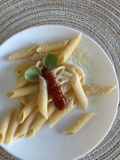 This is a close-up, overhead shot of a plate of penne pasta served with a dollop of red sauce and topped with grated cheese and basil leaves. The pasta is arranged artfully on a white plate, which is set on a circular, woven placemat. The background is slightly out of focus, but the texture of the placemat is clearly visible. The lighting is natural, creating soft shadows. There are no people visible in the image, and no indication of the specific location being in Гамово, Russia.