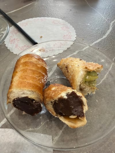 The image shows a clear glass plate containing three pastries. Two of the pastries are similar, golden-brown in color, with a ribbed exterior and a dark brown filling exposed at the broken end. One of these is intact, while the other is broken in half. The third pastry is also broken, showing a light-colored, possibly fruit-based, filling. The plate is placed on a gray marble-patterned surface, with a small white doily and a black straw visible to the left. The bottom of a glass is partially visible in the lower-left corner. There is no information in the image to indicate its location in Rosolini, Italy.