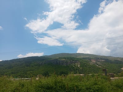 The image presents an outdoor scene in Zemen, Bulgaria, dominated by a verdant mountainous landscape under a partially cloudy blue sky. Two sets of parallel utility lines stretch horizontally across the upper mid-section of the frame. The hills are densely covered with dark green foliage, appearing heavily forested, with visible rocky outcrops and lighter, disturbed areas on the mid-right slope, suggesting a quarry or land development. In the mid-ground, a building with a prominent green roof and discernible lettering, likely spelling "ZEMEN," is visible behind the foreground vegetation. To its right, partially obscured industrial or abandoned structures with rusted components are present. The immediate foreground is characterized by a dense growth of unkempt green bushes and tall weeds. No individuals or specific human activities are depicted.