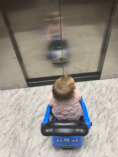 The image shows a young child from behind, sitting in a blue toy car with a black steering wheel. The child is wearing a pink quilted vest. The child and the toy car are positioned on a marble-patterned floor. Behind the child, there are two large, reflective stainless steel doors, which appear to be elevator doors. The reflection in the doors shows a blurry image of a person, likely the one taking the photo, and a blue object, possibly another toy or a stroller. The overall setting suggests an indoor environment, possibly a public space like a mall or an apartment building lobby. There is no indication of the time of day or weather conditions. The mood appears to be neutral, with no strong emotions conveyed. Visible text on the toy car reads "BABY CAR".