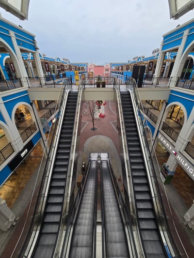 The image captures a multi-level interior of a shopping mall, likely an outlet mall, with a symmetrical design. Two sets of escalators, one on the left and one on the right, dominate the foreground, extending upwards towards the upper levels. The architecture features blue and white facades with arched openings leading to various storefronts. Balconies with black railings overlook the central atrium. The floor is paved with brick-like tiles in the central walkway, and the shops are visible through glass windows. Signs for brands like "Tommy Hilfiger" are visible. The lighting suggests it is daytime, but the sky appears overcast. The scene is devoid of people, presenting a quiet and empty shopping environment. The location is identified as the Fuyang Outlet Mall in Fuyang, China.