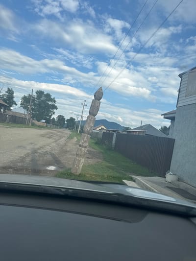 An outdoor, eye-level, medium shot captured from inside a vehicle shows a village scene in Yabogan, Russia. The foreground is dominated by the interior of a car, with the dashboard and windshield frame visible. Beyond the vehicle, a dirt road winds through the village, flanked by wooden fences and houses.

A prominent feature in the right-center of the image is a leaning wooden pole, constructed from stacked logs, which appears to be supporting overhead electrical wires. The pole is weathered and shows signs of age. In the background, a few trees and distant mountains under a partially cloudy blue sky complete the rural landscape. The overall impression is that of a quiet, perhaps remote, village on a bright day.