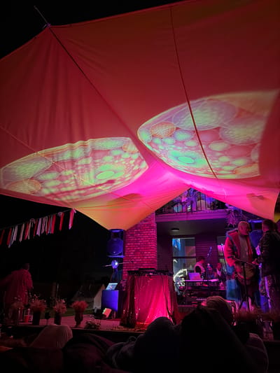 The image depicts an outdoor nighttime event with a stage set up. A large red canopy stretches overhead, illuminated with projected light patterns that resemble cellular structures. The canopy is partially supported by brick walls. A band or musical group is performing on the stage, with several individuals visible playing instruments and operating equipment such as a keyboard and DJ decks. The stage is draped with a shimmering pink fabric. In the foreground, an audience is seated, appearing to relax on the ground or low seating. Decorative elements like streamers and vases of dried flowers are present. The overall atmosphere suggests a music festival or outdoor concert. The location is stated to be in Shebanovo, Russia.