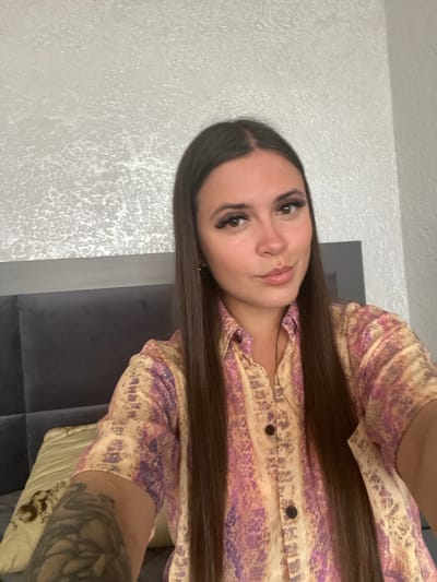 A woman with long, dark hair, wearing a collared, short-sleeved shirt with a patterned design in shades of pink and yellow, is captured in a close-up, upper-body selfie. Her head is slightly tilted, and she has on false eyelashes. She is positioned in front of a textured, light-colored wall and a dark-colored headboard, suggesting she is indoors, likely in a bedroom. A pillow with a tan and black pattern is visible to her left. There is a tattoo visible on her left arm. The scene does not provide any specific information to determine a location in the city of Chalda, Russia.