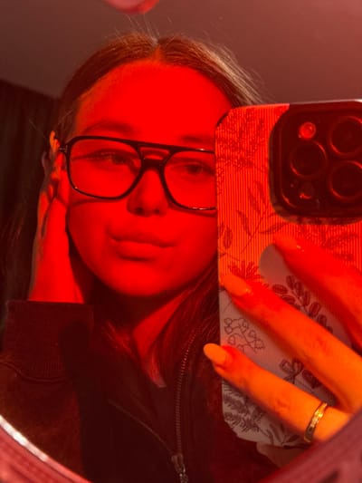 A female subject, located in Prikubanskiy, Russia, is depicted in a mirror selfie under red illumination. She is wearing dark-rimmed, rectangular glasses and a dark, zippered jacket. Her right hand is positioned against the side of her head, near her temple. Her left hand, featuring nails with light orange polish and a gold-colored ring on the ring finger, holds a smartphone with a floral-patterned case and a visible multi-lens camera module. The subject's reflection shows her facing forward, looking into the mirror. The immediate background is indistinct due to the lighting.